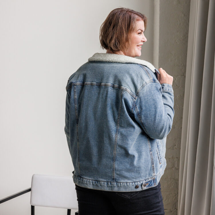 Middle aged brown haired female model wearing a denim sherpa lined jacket with aged-brass buttons, and black pants. denimandpatches