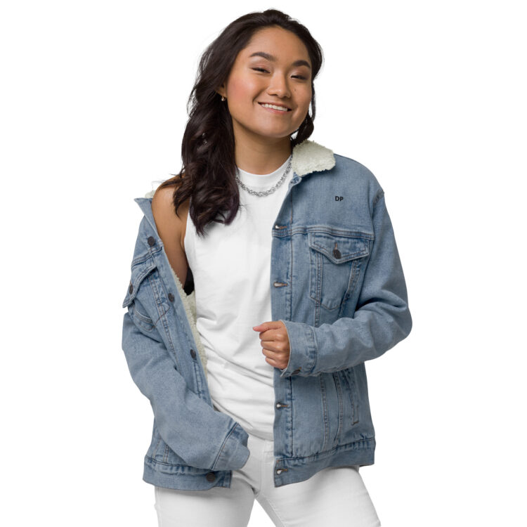 Young female model featuring white clothes underneath the light blue denim sherpa lined jacket. The jacket featuring aged-brass buttons to close the jacket and a frocket being held closed by the same button. The letters DP written in small/bold black font above the front left frocket. denimandpatches