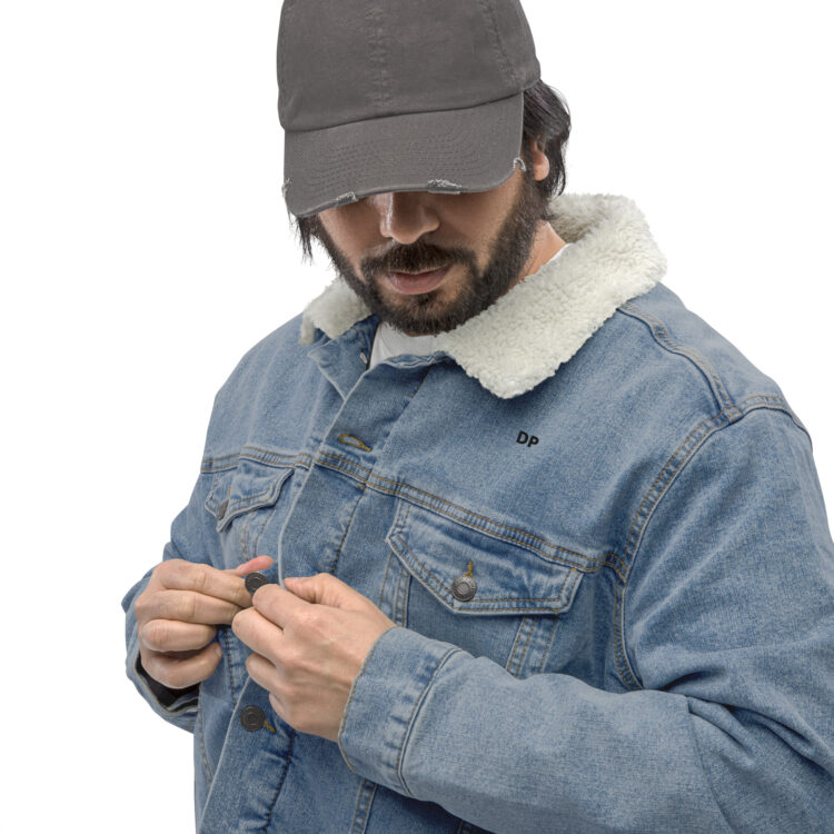 Side/front shot of a brunette male model. In mid closing of our light blue sherpa-lined denim jacke wearing a black baseball cap. The model is using the antique brass buttons to close the jacket, the buttons are also featured closing the 2 front chest pockets. Above the front left pocket the letters "DP" are printed in bold black lettering. denimandpatches