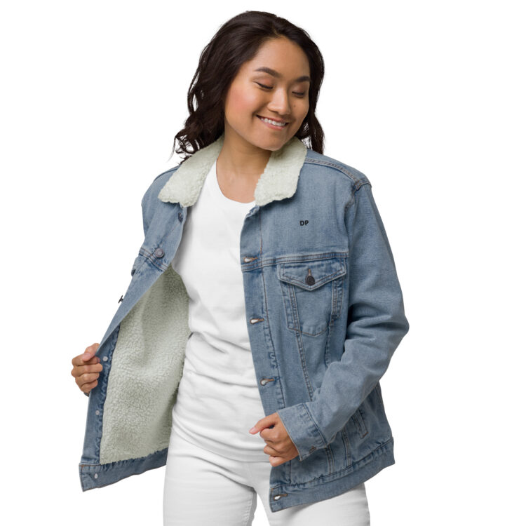 Young female model wearing white clothes underneath the light blue denim sherpa lined jacket. The jacket featuring aged-brass buttons to close the jacket and a frocket being held closed by the same button. The letters DP written in small/bold black font above the front left frocket. denimandpatches