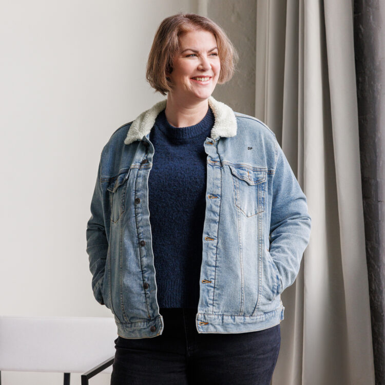 Middle aged female model wearing a denim sherpa jacket. The jacket featuring a soft sherpa lining with 2 frockets that are closed with a flap and a aged-brass button. Also features brass buttons to close the jacket to. denimandpatches