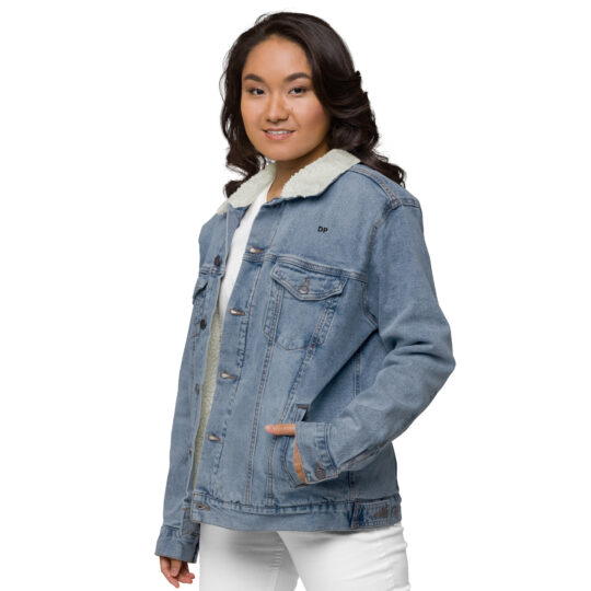 Young female model wearing white clothes underneath the light blue denim sherpa lined jacket. The jacket featuring aged-brass buttons to close the jacket and a frocket being held closed by the same button. Her left hand is using the hand pockets at the bottom of the jacket. The letters DP written in small/bold black font above the front left frocket. denimandpatches