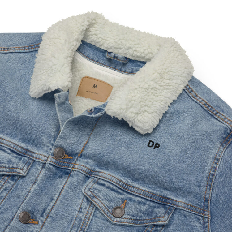 Image tries to highlight our light blue sherpa denim jacket collar. It again shows the fluffy white sherpa lining that can be removed. With a loop to hang the jacket built into the collar with a brown "leather" tag underneath said loop.The jacket also features "DP" written in small/bold black letters. above the front left frocket. that is held closed by a aged-brass button. The jacket is also being held closed by the same brass buttons. denimandpatches