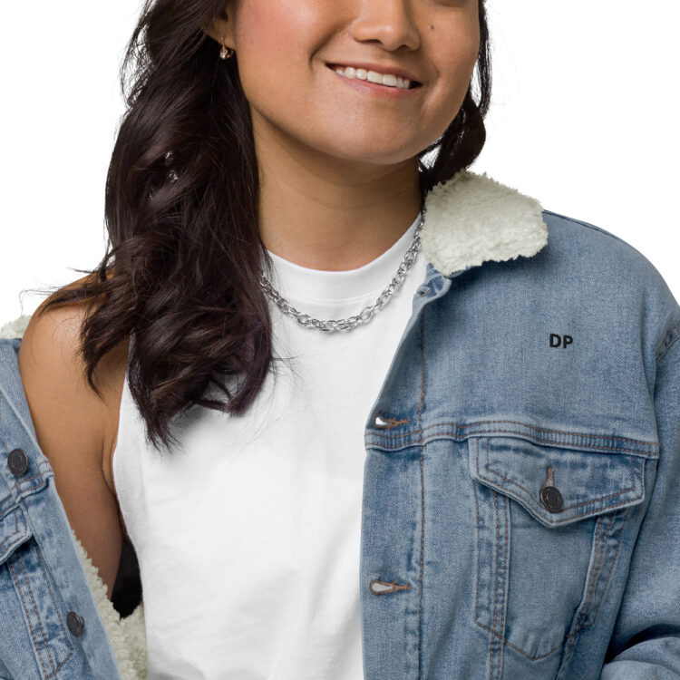 Zoomed in image of young female model wearing white clothes underneath the light blue denim sherpa lined jacket. The jacket featuring aged-brass buttons to close the jacket and a frocket being held closed by the same button. The letters DP written in small/bold black font above the front left frocket. denimandpatches