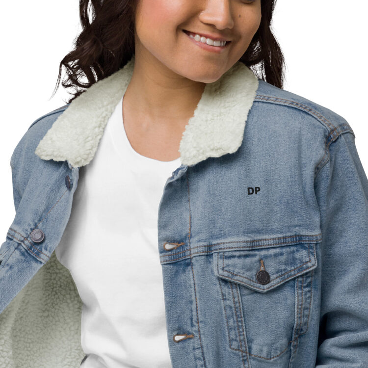 Zoomed in image of young female model wearing white clothes underneath the light blue denim sherpa lined jacket. The jacket featuring aged-brass buttons to close the jacket and a frocket being held closed by the same button. The letters DP written in small/bold black font above the front left frocket. denimandpatches